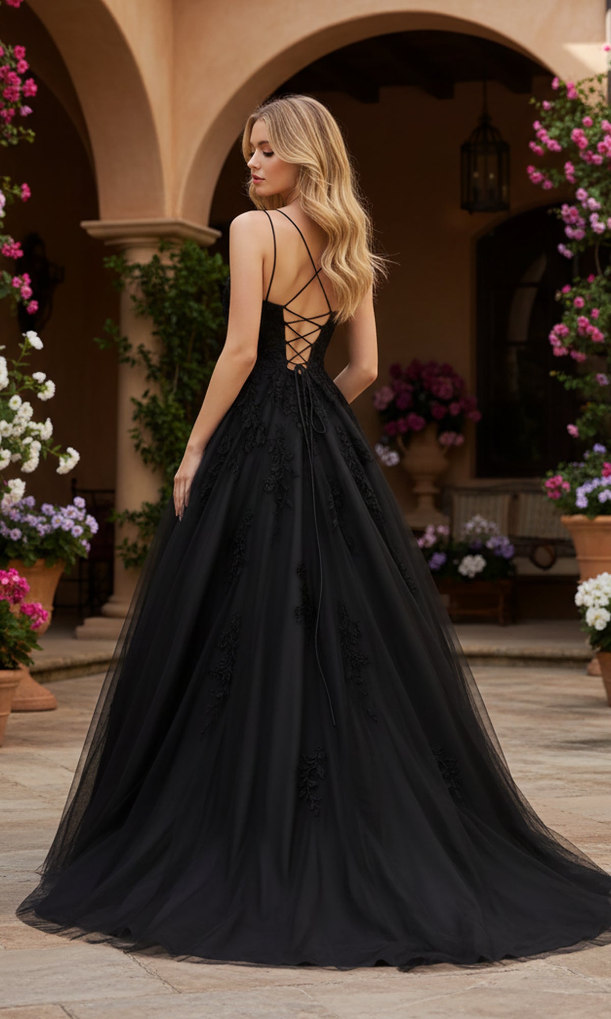 A woman wears the PromGirl Exclusive 250228 long black lace prom dress with an adjustable lace-up back, standing outdoors on stone pavement among potted blooming plants and arched architecture.